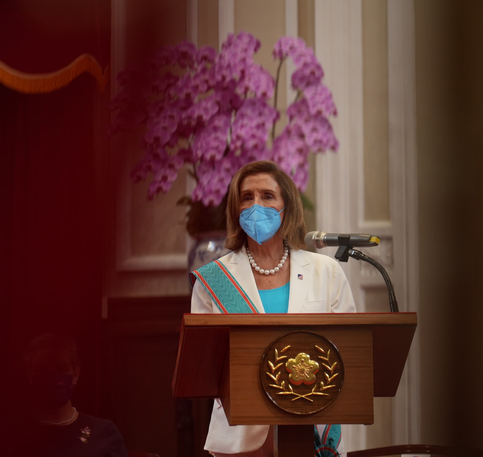 Pelosi in a face mask, delivering a speech at the podium