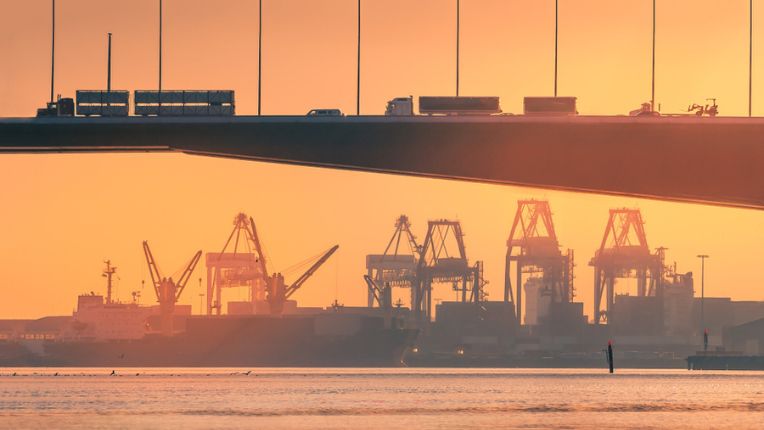 A road bridge with trucks, cars and a fork lift truck is in silhouette over an industrial skyline