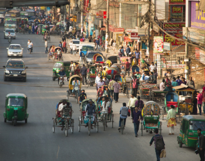 A busy street in Dhaka with cars, rickshaws, and pedestrians amid shops and roadside vendors in an urban area during daylight.